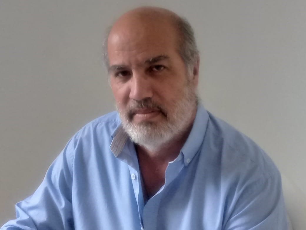 Homem calvo com barba grisalha curta vestindo camisa social azul clara em fundo branco.