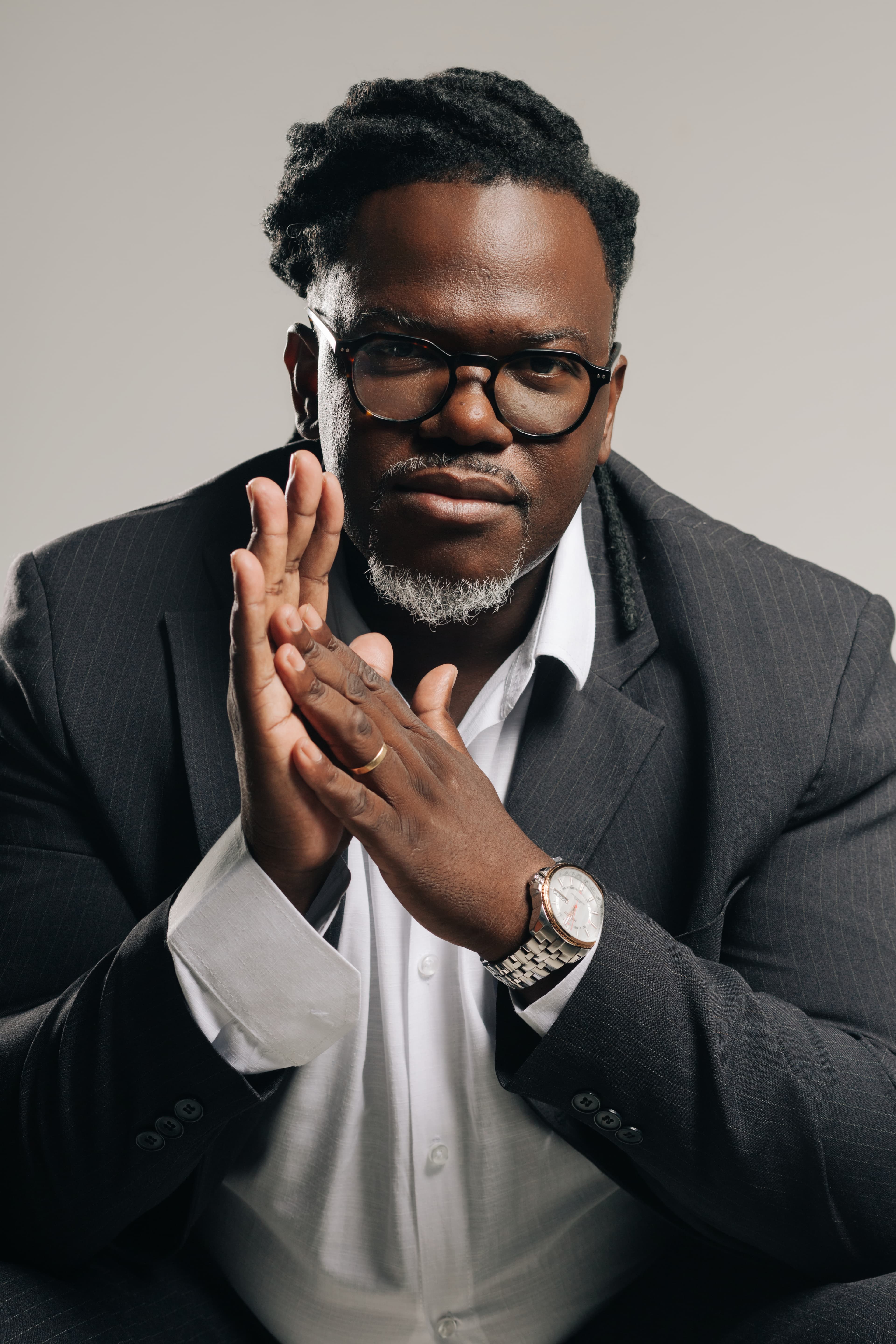 Homem negro de óculos e terno, com as mãos unidas em um retrato profissional.