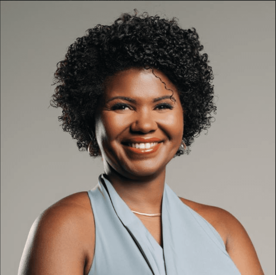 Mulher negra sorridente com cabelo crespo curto, vestindo blusa azul clara e colar dourado.