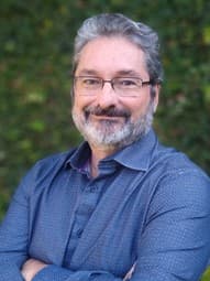 Homem sorridente com cabelos grisalhos, barba e óculos, vestindo camisa azul em fundo verde.